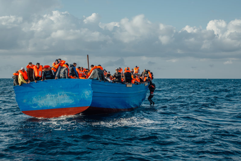Bei einem stark überbesetzten Holzboot hält sich rechts ein Mann an der Reling fest, um nicht ins Meer zu fallen