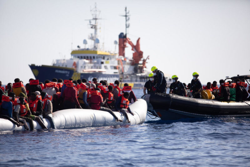 Im Vordergrund ein überbesetztes Schlauchboot links sowie ein schnelles Rettungsboot rechts. Im Hintergrund das Rettungsschiff Humanity 1.