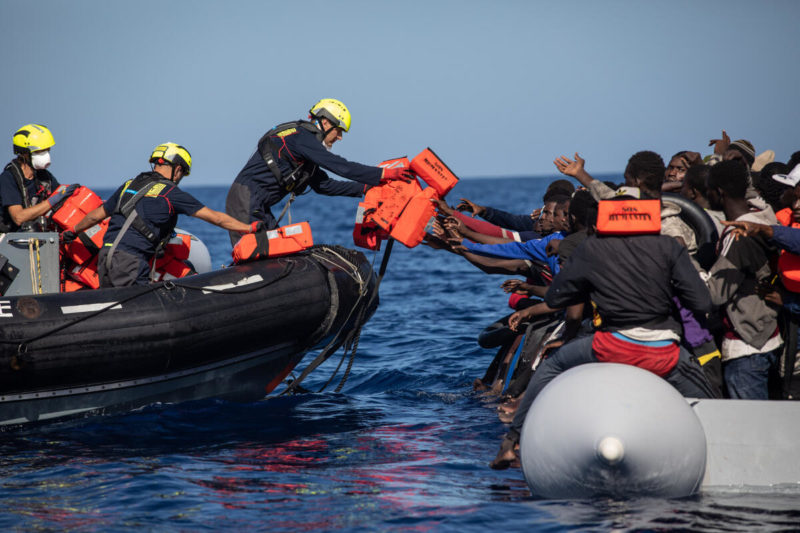 Bei einem Rettungseinsatz im Mittelmeer reicht ein Rettungsteammitglied eine Rettungsweste an einen Menschen in einem überbesetzten Schlauchboot.