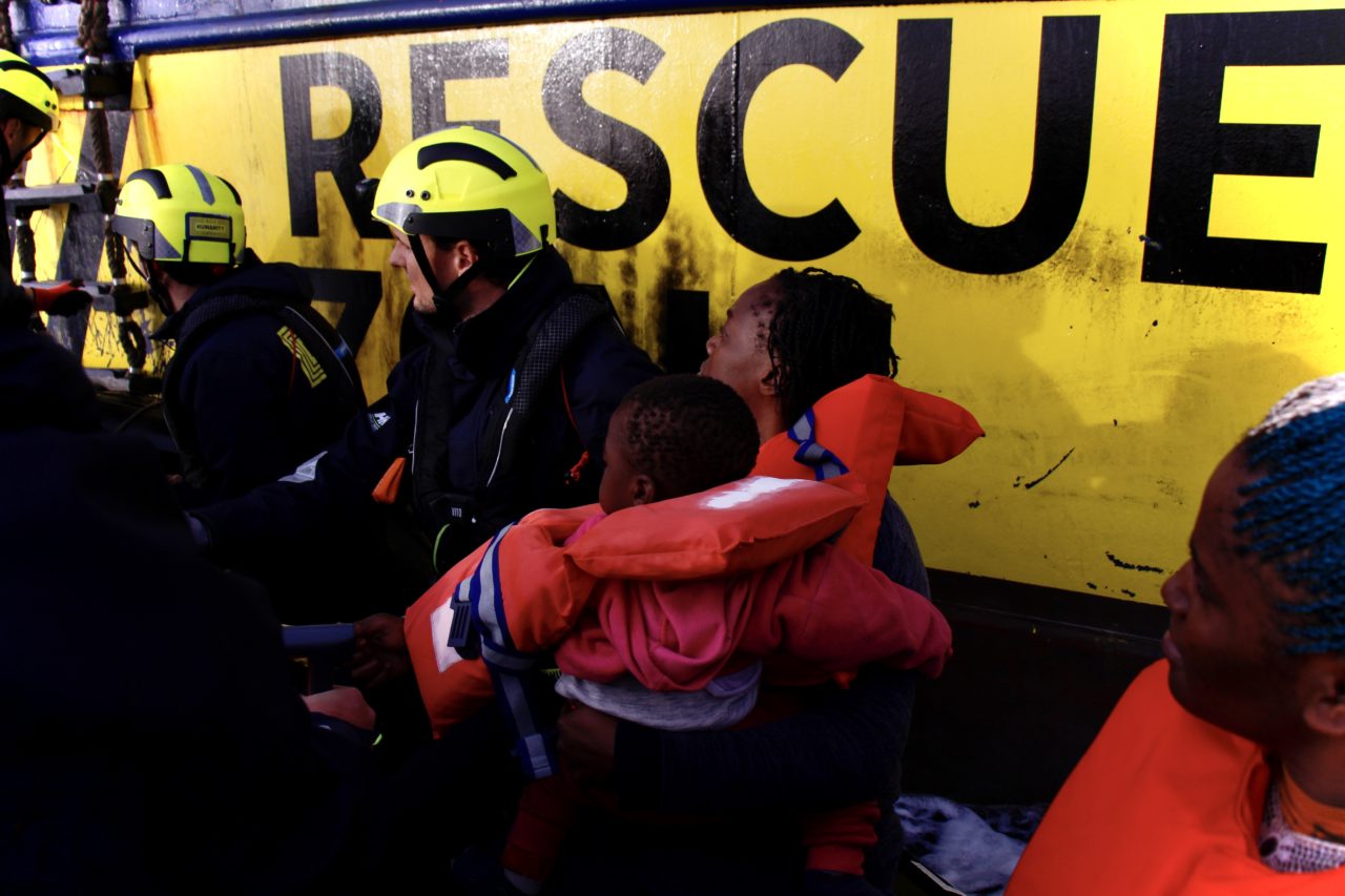 Vor der Bordwand der Humanity 1 sind links Frauen und Kinder in Rettungswesten, rechts Mitglieder des Rettungsteams zu sehen.