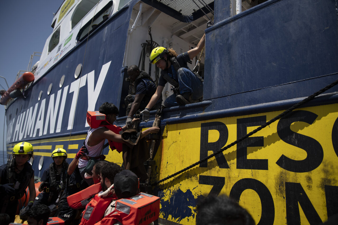 EIn Crewmitglied hilft einem Geretteten von einem Rettungsboot in die blau-gelbe Humanity 1 (Rettungsschiff von SOS Humanity).