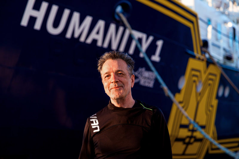 Tom (Software-Entwickler für Registrierungsprogramm an Bord) lächelnd vor dem blau-gelben Rettungsschiff Humanity 1.