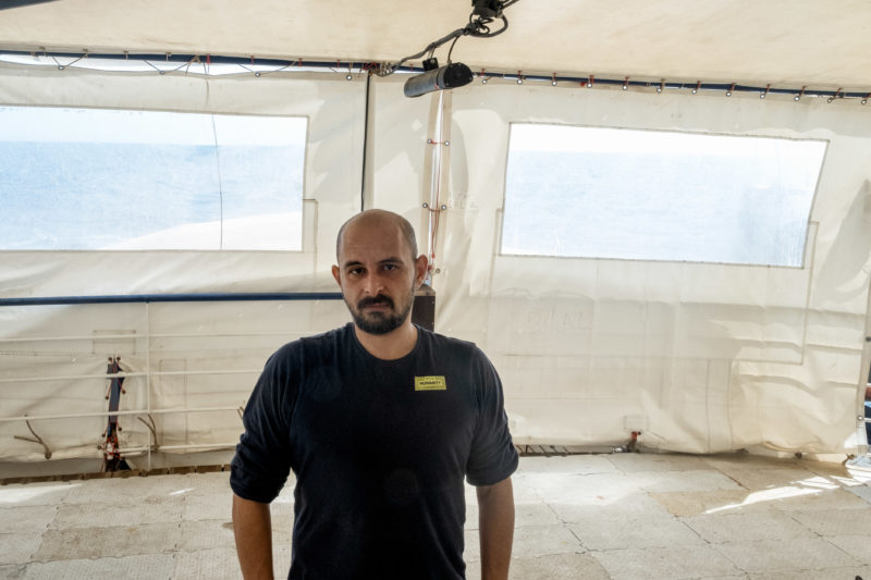 Halbtotale von Fares mit blauen SOS Humanity T-Shirt an Bord der Humanity 1 mit weißem Hintergrund und Ausblick auf den blauen Himmel und das Meer
