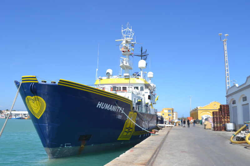 Humanity 1 in den SOS Humanity-Farben gelb und blau im Hafen von Siracusa mit blauem Himmel und der Stadt im Hintergrund.