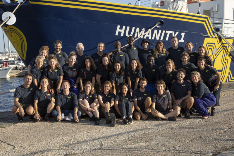 Crew von SOS Humanity vor dem Rettungsschiff Humanity 1 mit blauen T-Shirts.