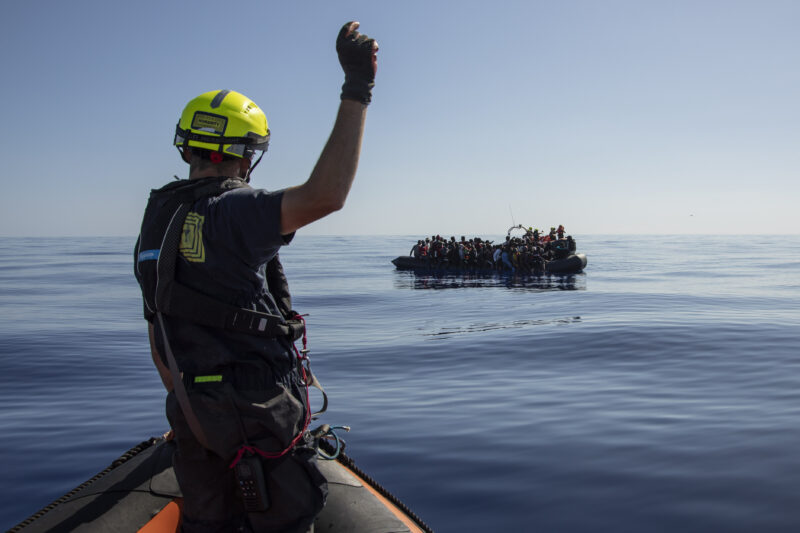 Crewmitglied von SOS Humanity von hinten mit gelbem Helm und erhobener Hand auf einem RHIB, im Hintergrund ein Boot mit Überlebenden.