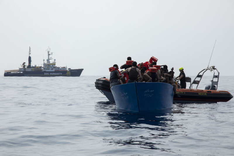 Blaues Boot auf dem Mittelmeer mit Flüchtlingen an Bord, dahinter SOS Humanity Rettungsboote und die Humanity 1.