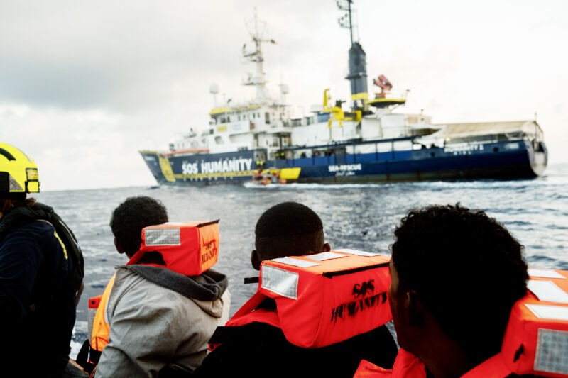 Gerettete von hinten, die rote Rettungswesten tragen und gerade an Bord der Humanity 1 gebracht werden.