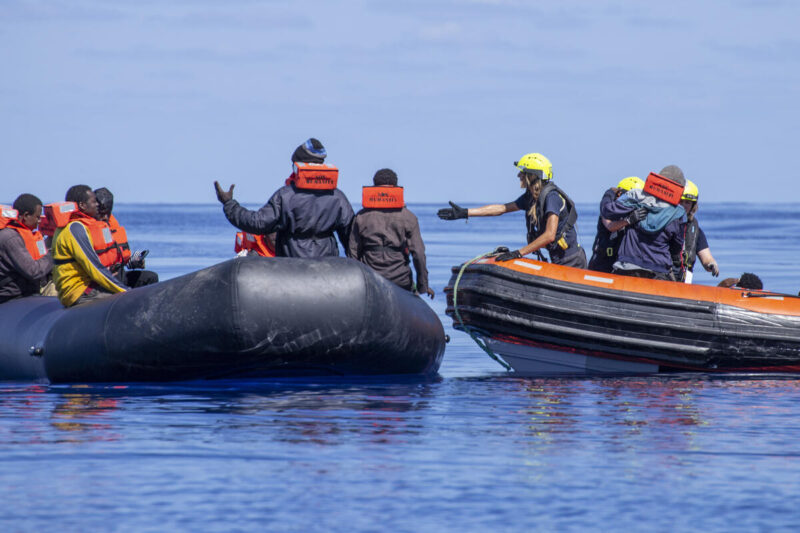 RHIB von SOS Humanity rechts und Schlauchboot mit Geflüchteten links. Retterin hilft den Menschen mit ausgestreckter Hand auf das Schnellboot.