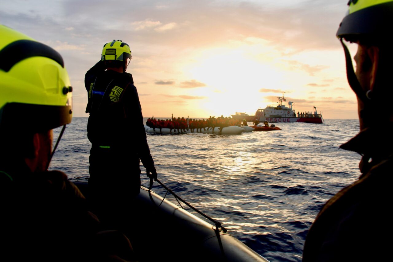 Drei Crewmitglieder mit gelben Helmen Blicken auf das Meer und den Sonnenunter- oder -aufgang im Hintergrund.