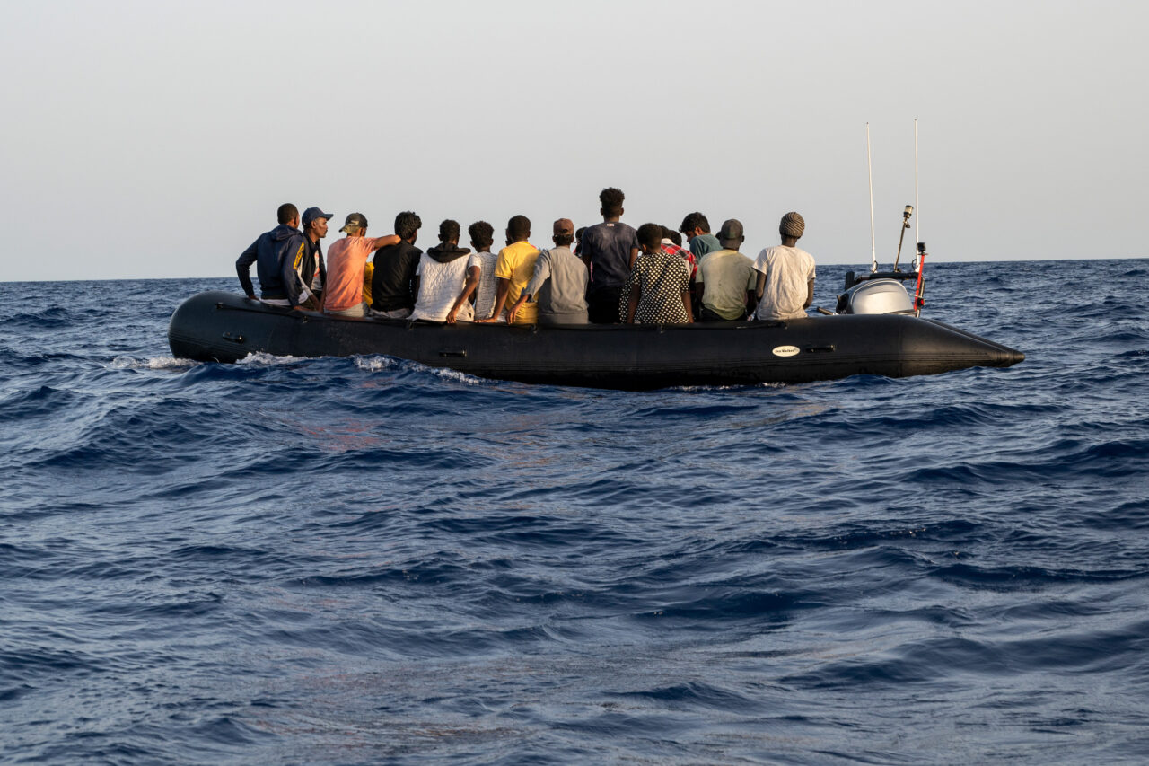 Flüchtlingsboot mit mehreren Personen von hinten auf dem zentralen Mittelmeer.