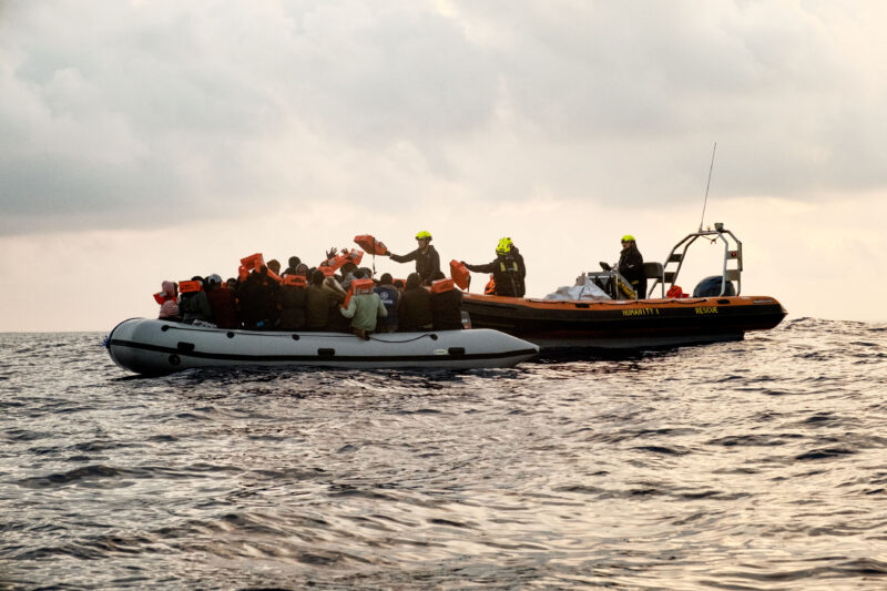 Links Schlauchboot mit Flüchtlingen, rechts RHIB mit Crewmitgliedern, die Rettungswesten austeilen.