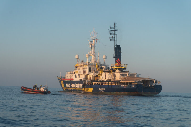 Die Humanity 1 (Rettungsschiff von SOS Humanity) in gelb und blau, daneben das schnelle Rettungsboot RHIB und im Hintergrund die Morgen- oder Abendsonne auf dem Mittelmeer.