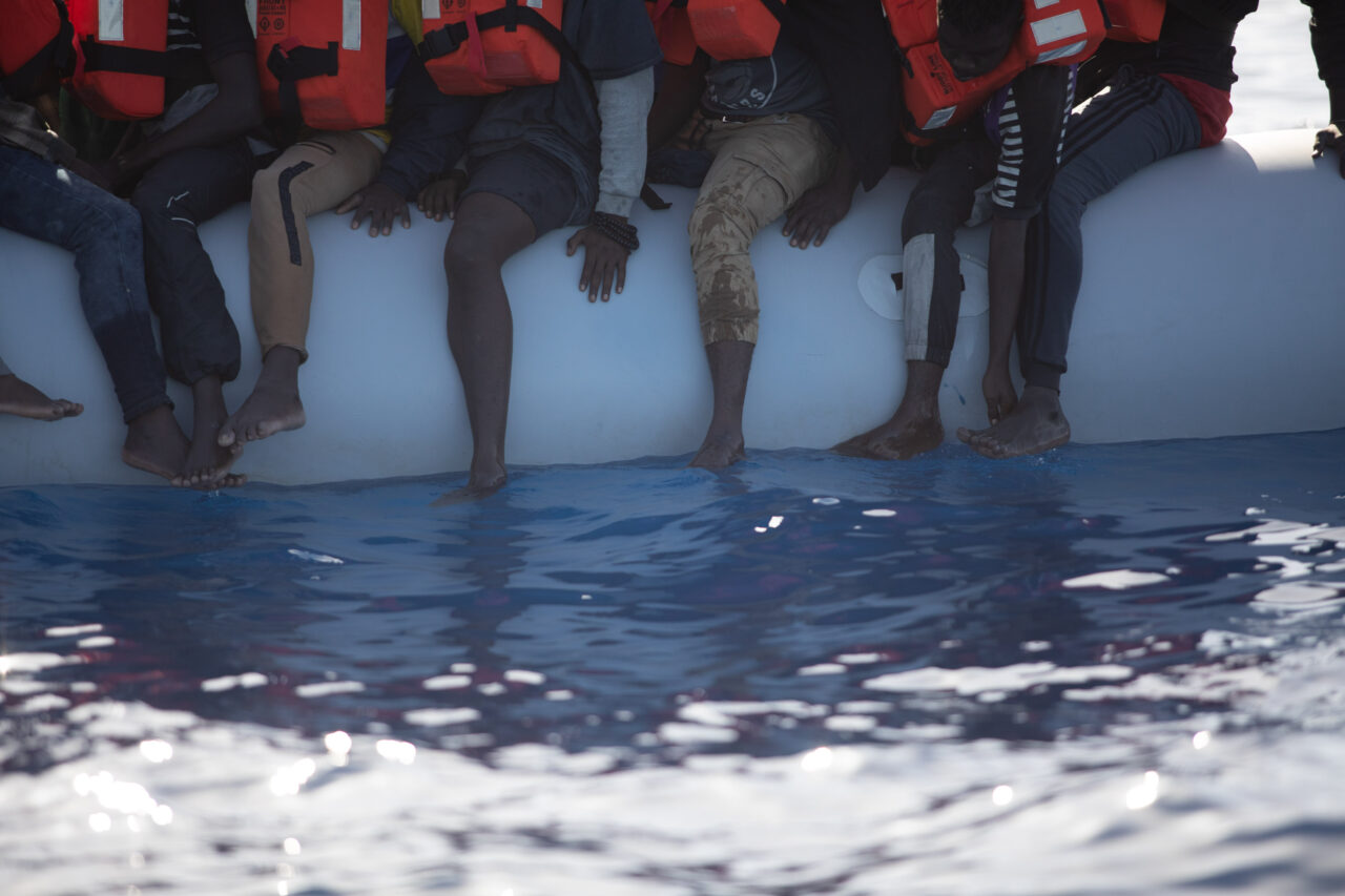 Beine und Füße von Menschen, die auf einem Schlauchboot sitzen, und auf dem Vordergrund Wasser.