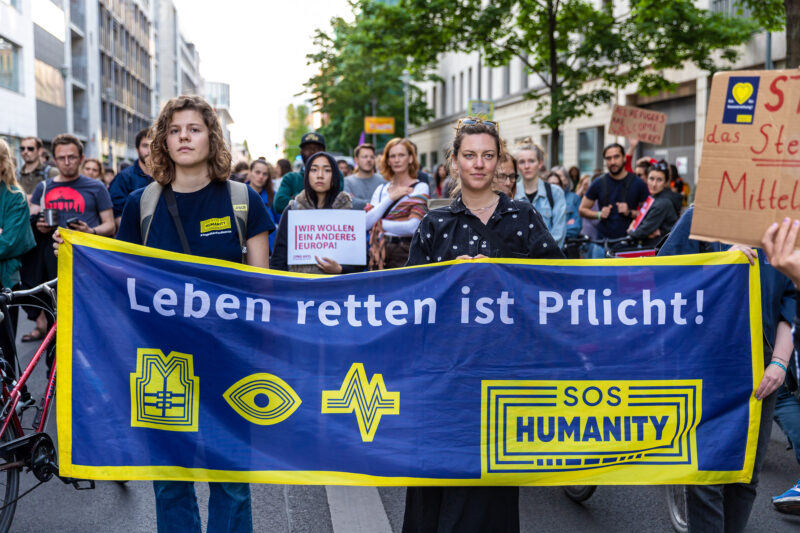 Teilnahme von SOS Humanity bei einer Demo. Zwei Personen halten ein blau-gelbes Banner mit der Aufschrift "Leben retten ist Pflicht"