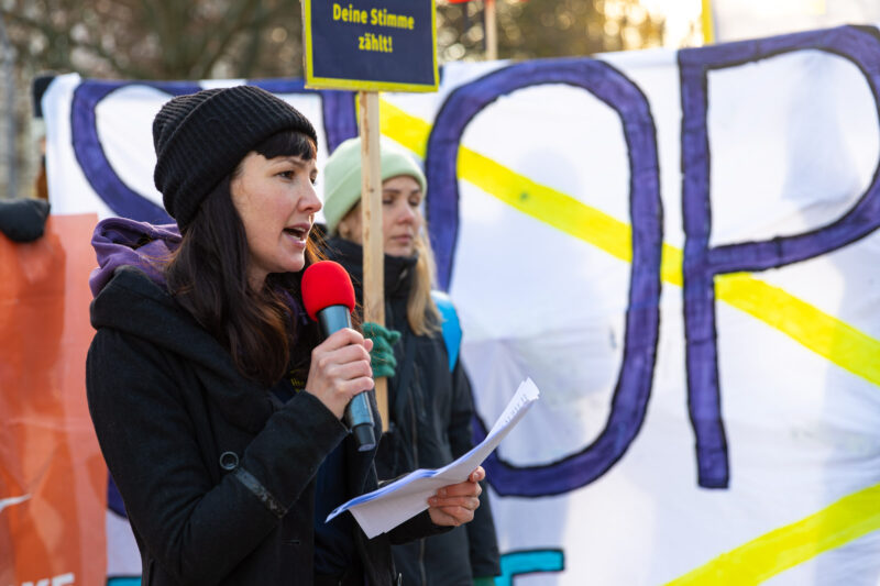 Lisa aus dem Vorstand von SOS Humanity mit einer Jacke und schwarzer Mütze und einem roten Mikro bei der GEAS Demo in Berlin.