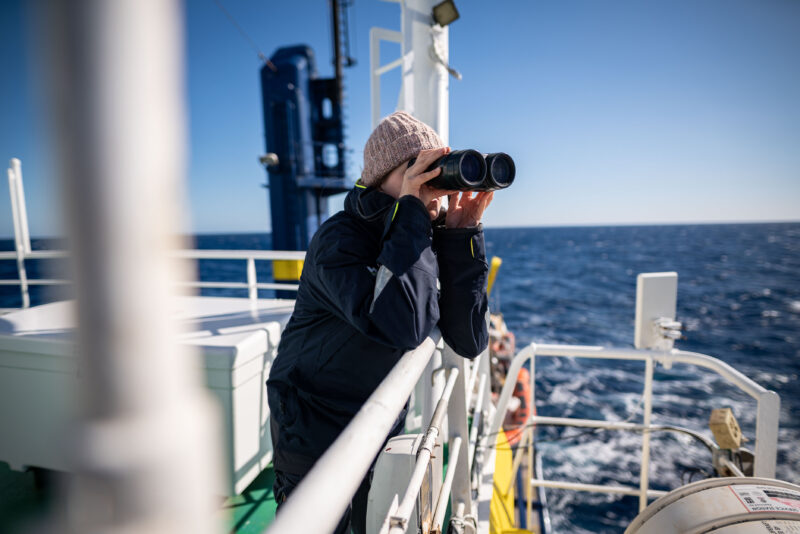Crewmitglied beim Look-Out an Bord der Humanity 1 mit Ferngläsern und Mütze.