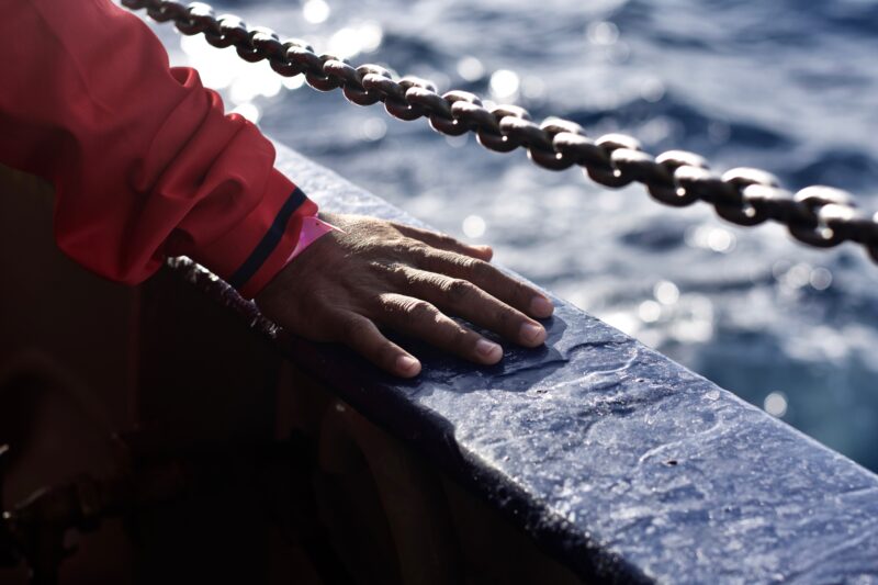 Hand eines Geretteten auf dem Reling der Humanity 1 mit einer Kette und dem Meer im Hintergrund.
