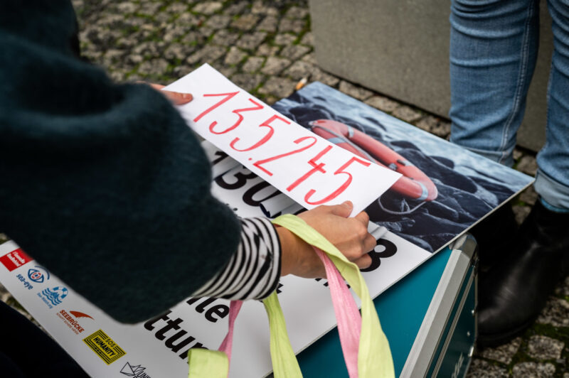 Hände kleben ein Blatt Papier mit der Zahl 135245 auf ein Plakat für eine Petitionsübergabe von SOS Humanity vor dem Bundestag in Berlin.