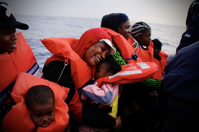 Eine Frau trägt eine Schwimmweste und hält weinend ihr Baby, auch mit Schwimmweste, in der Hand. Die beiden befinden sich auf einem Rettungsboot im Mittelmeer, zusammen mit weiteren Geretteten.