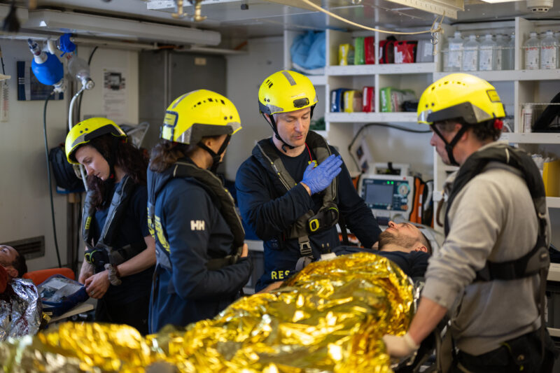 Drei Crewmitglieder stehen um eine Person, die unter einer goldenen Wärmedecke auf einer Liege im Hospital der Humanity 1 liegt.