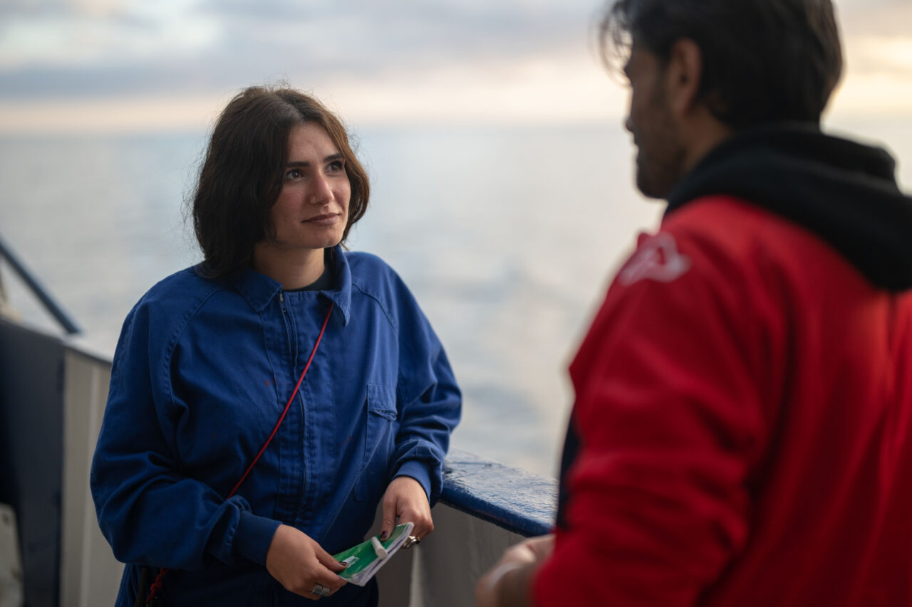Eine Frau in blauem Overall steht einem Mann in rotem Pullover gegenüber. Sie scheint ihm aufmerksam zuzuhören.