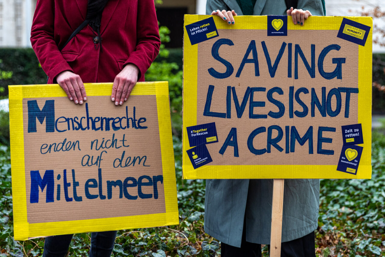 Zwei Demo-Schilder mit gelben Rahmen, auf denen "Menschenrechte enden nicht auf dem Mittelmeer" und "Saving Lives is not a crime" steht.