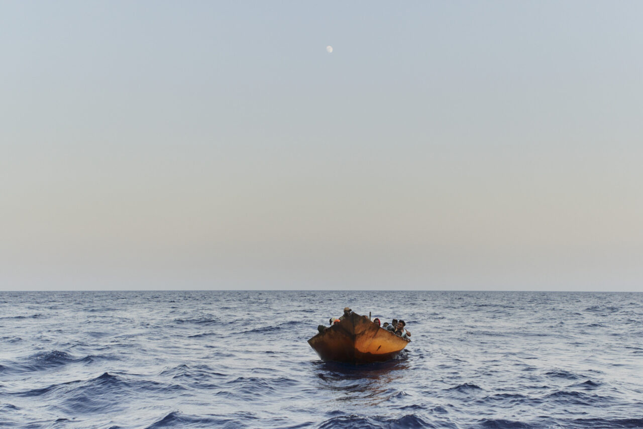Auf dem Mittelmeer unter blauem Himmel treibt ein rötliches Holzboot mit mehreren Menschen an BOrd.