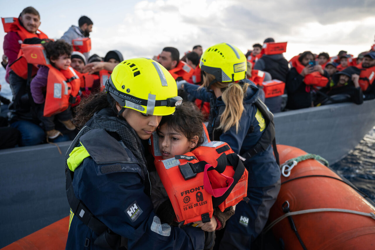 Ein kleines Kind mit Rettungsweste wird von einer Seenotretterin von SOS Humanity auf ein Boot gebracht.