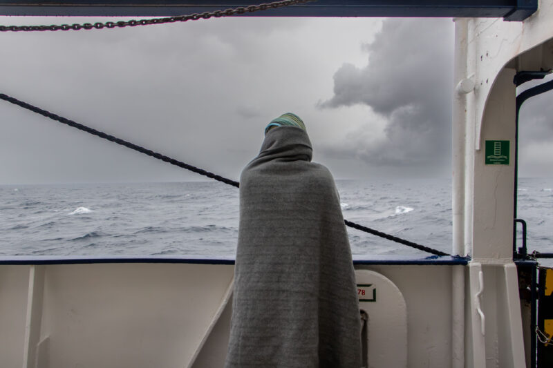 Überlebender auf der Humanity 1 ist eine graue Decke gehüllt und blickt auf das stürmische Mittelmeer.
