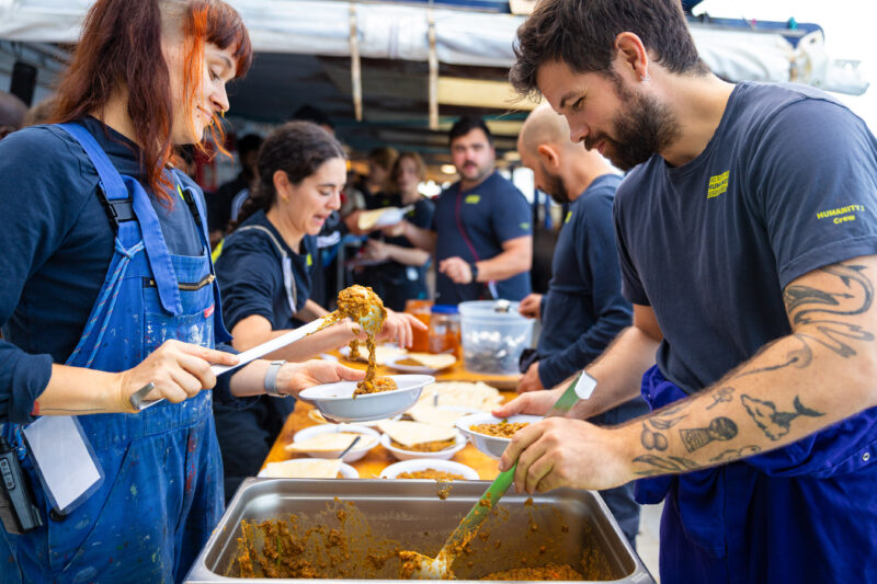 Zwei Crewmitglieder mit blauen SOS-Humanity T-Shirts geben Essen an Bord der Humanity 1 aus.