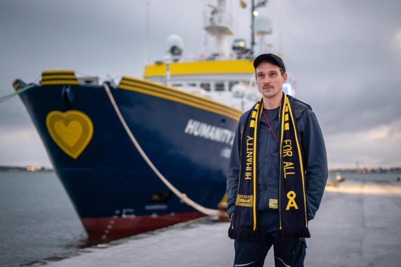 Daniel Sträßer, ein deutscher Schauspieler, steht vor der Humanity 1 und trägt einen SOS Humanity Schal mit der Aufschrift Humanity 4 All.