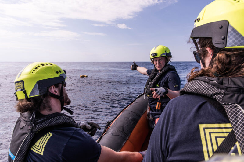 Drei der SOS Humanity Crew mit gelben Helmen auf einem Schlauchboot auf dem Meer.