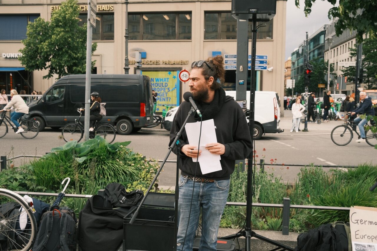 Speaker is standing in front of a microphone in front of the European House.